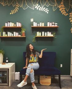 a woman sitting on a blue chair in front of shelves filled with bottles and plants