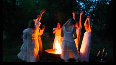 four women dressed in white dancing around a fire pit at night with their arms raised