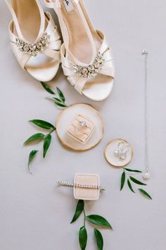 wedding shoes and jewelry laid out on a white tablecloth with green leaves around them