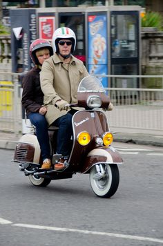 a man and woman riding on the back of a scooter down a street