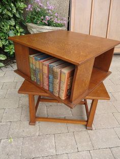 a small wooden table with books in it on the ground next to some bushes and flowers