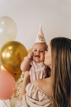 a woman holding a baby wearing a party hat and kissing her face with balloons in the background