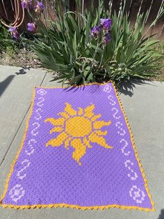 a purple and yellow crocheted blanket sitting on top of a sidewalk next to flowers