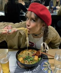 a woman is eating noodles with chopsticks at a restaurant while wearing a red hat