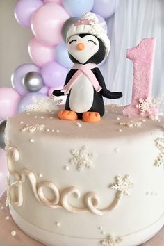 a birthday cake with a penguin on top and balloons in the background for a first birthday