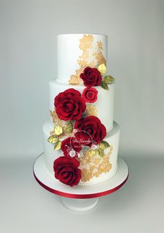 a three tiered white cake with red roses on the side and gold leaf decorations