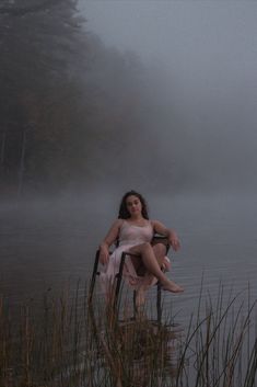 a woman sitting in a chair on the edge of a body of water with fog