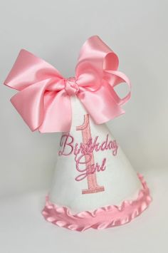 a white birthday hat with pink ribbon and a number 1 on the side, decorated with a large pink bow