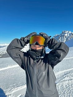 a man wearing ski goggles standing on top of a snow covered slope with his hands behind his head