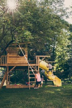 two children playing on a wooden play set in the grass under a tree with a yellow slide