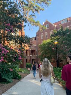 people walking down a sidewalk in front of a building with pink flowers on the trees