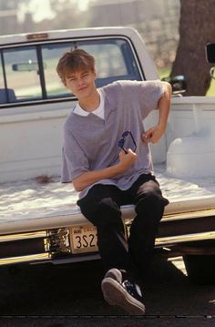 a young man sitting on the back of a truck in front of a parked car