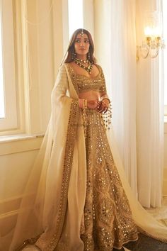 a woman standing in front of a window wearing a gold and white lehenga