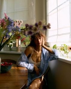 a woman sitting at a table with flowers in the window sill behind her and looking off into the distance