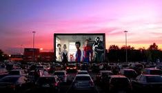 an outdoor movie screen in the middle of a parking lot with cars and people watching it