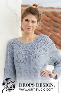 a woman standing next to a white wall wearing a blue knitted sweater and smiling