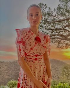 a woman in a red and white dress standing next to a tree with the sun setting behind her