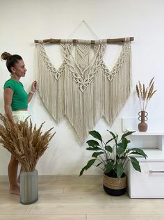 a woman standing in front of a macrame wall hanging on a wall next to two potted plants