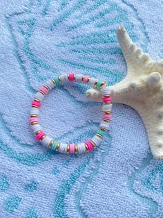 a white starfish with pink, green and gold beads on a blue towel next to a seashell