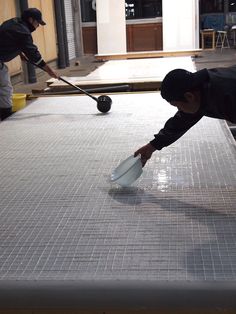 two men are cleaning the floor in an industrial area, one is using a broom to clean the tile