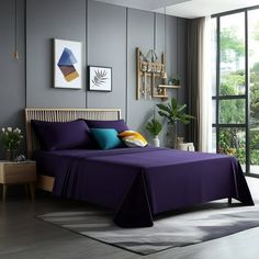 a bed with purple sheets and blue pillows in a room next to a large window