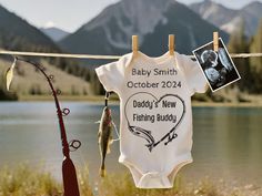 a baby bodysuit hanging on a clothes line next to a lake and fishing rod