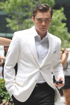 a man in a white suit and tie walking down the street with his cell phone