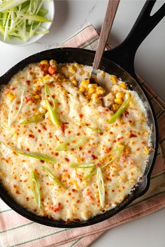 a skillet filled with corn and cheese on top of a towel next to a spoon