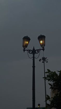 two street lamps are lit up in the dark sky above some trees and buildings on a cloudy day