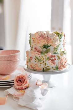 a three layer cake with pink flowers on it and plates around the cake, ready to be cut