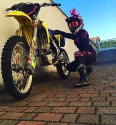 a person sitting on the ground next to a yellow dirt bike with pink hair and spikes