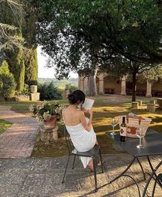 a woman sitting at a table reading a book and drinking wine in the back yard