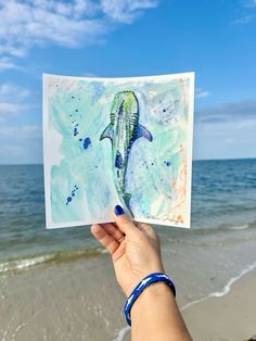 someone holding up a card with a painting of a dolphin on the beach in front of the ocean