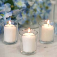 three white candles sitting on top of a table next to flowers and vases with blue hydrangeas in the background