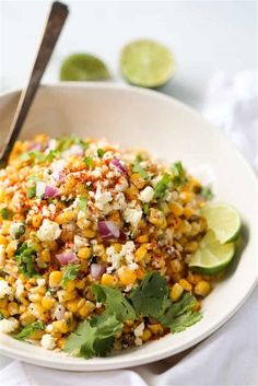 a white bowl filled with corn salad and topped with cilantro, lime wedges