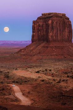 a full moon is seen over the desert with a car driving on it's side