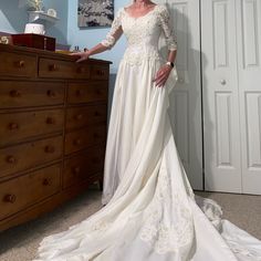 a woman standing in front of a dresser wearing a wedding dress with sleeves on it