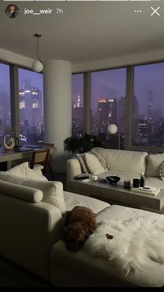 a living room filled with white furniture and large windows overlooking the city at night time