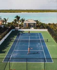 two people playing tennis on a blue and green court near the water with palm trees