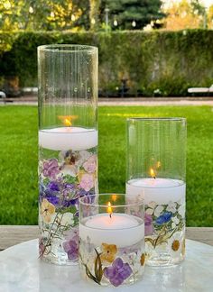 three glass vases filled with flowers and lit candles on a table in front of a grassy area