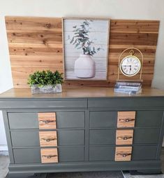 a gray dresser with wooden drawers and a clock on top, next to a plant