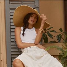 a woman wearing a large hat sitting on top of a wooden bench next to a plant