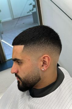 A profile image of a man with a sharp, high fade haircut. His hair is dark and closely cropped on top, with a gradual fade down to the skin on the sides. He has a neatly trimmed beard and is wearing a diamond stud earring in his left ear. The man is seated in a barber chair, wearing a white barber cape. The background shows a modern, clean barbershop setting with a window and some greenery outside. The image is captioned "2024's Hottest Men's Fade Haircuts Short & Medium Styles" at the bottom. Fade With Braids Men, High Drop Fade, Short Haircut Men, Boosie Fade, Best Fade Haircuts