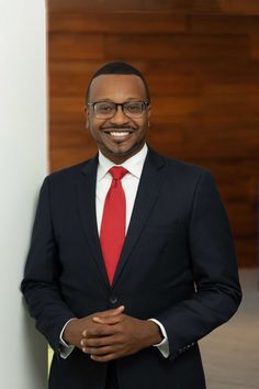 a man in a suit and red tie standing next to a white wall with wood paneling