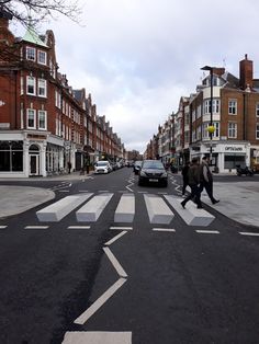 two people crossing the street in front of cars and buildings on either side of an arrow painted crosswalk