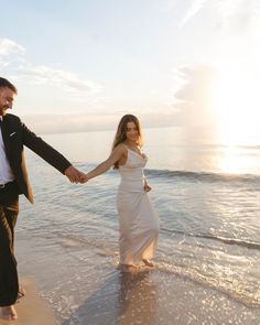 a man and woman holding hands walking on the beach at sunset with waves coming in