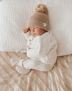 a baby laying on top of a bed wearing a hat and white sweater with buttons