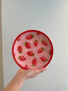 a hand holding a red and white plate with strawberries printed on the bottom, in front of a white wall