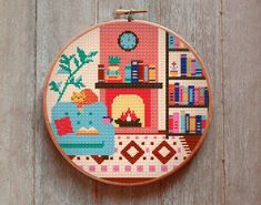 a cross stitch pattern on a hoop hanging from a wooden wall with a fireplace and bookshelf in the background