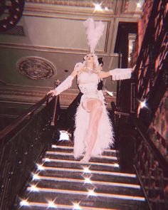 a woman dressed in white is walking down the stairs with feathers on her head and legs